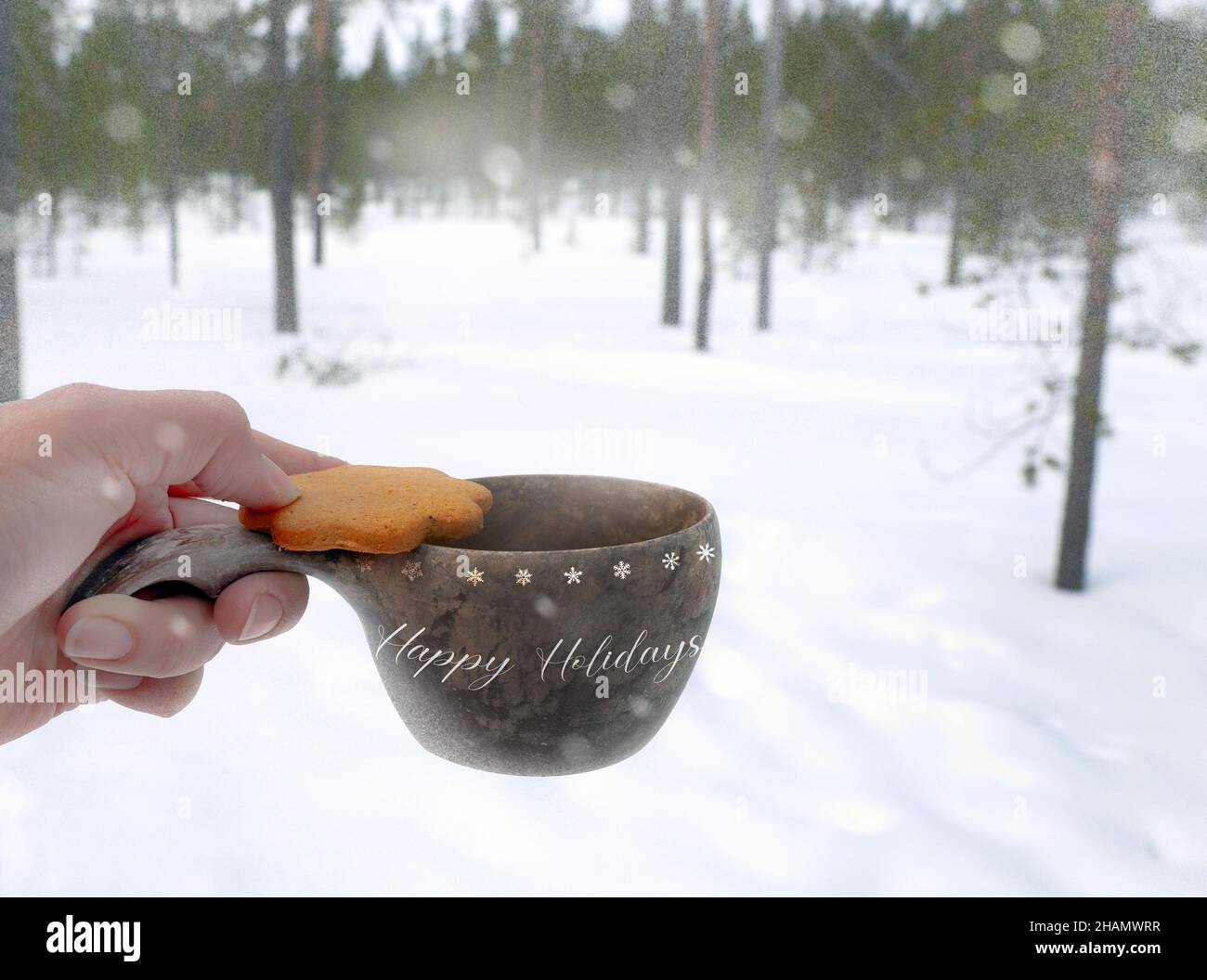 una mano che tiene biscotti con bevanda calda in una tazza di legno all'esterno con la vista della foresta di pini sotto la copertura delle feste felici di nevicatura bianche Foto Stock