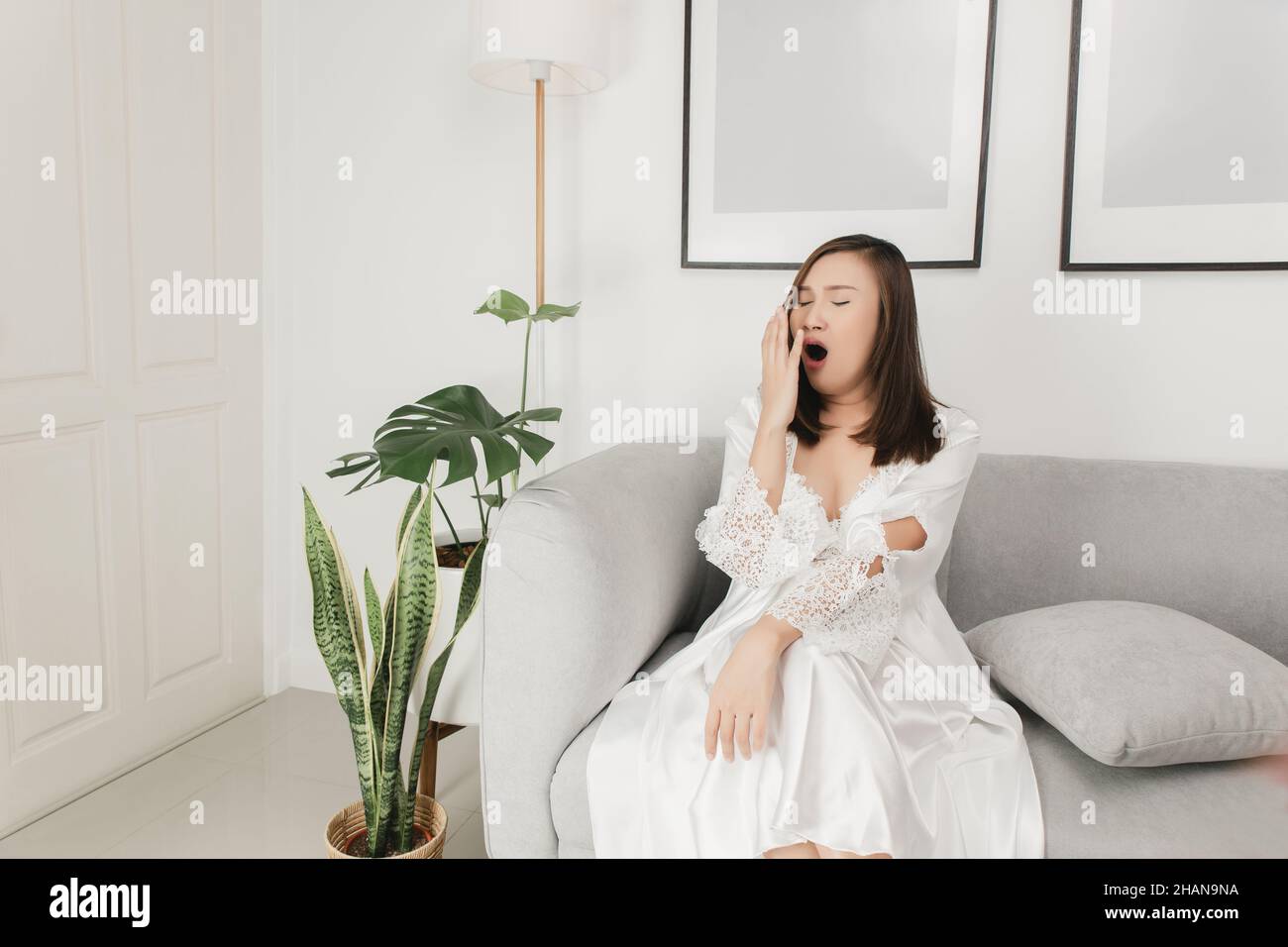 Donna thailandese in un nightgown satinato e vestito di abito bianco che si yawning sul divano nel soggiorno di notte Foto Stock