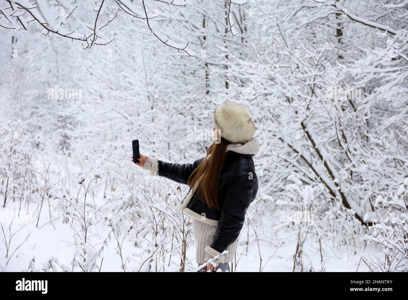 Ragazza in cappotto nero e cappello in pelliccia con selfie su smartphone fotocamera sullo sfondo della natura neve nella foresta invernale. Alberi dopo la nevicata Foto Stock
