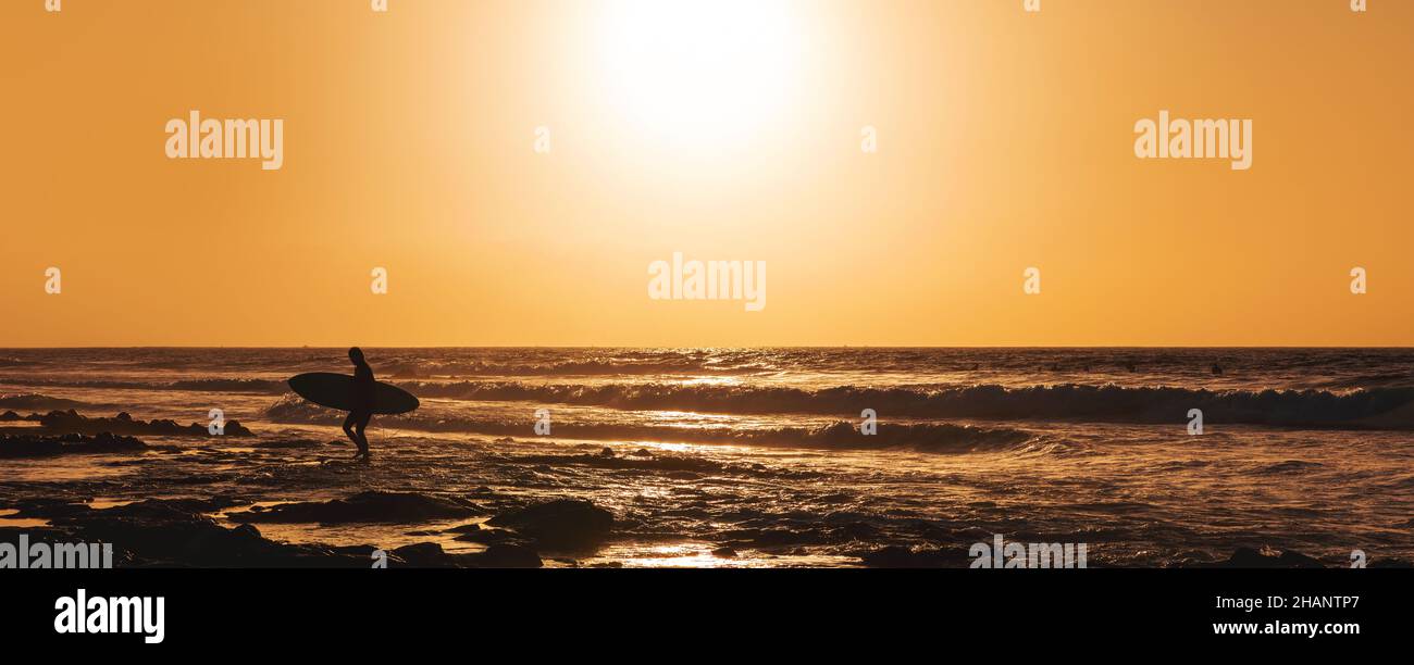 silhouette di surfista con tavola da surf che esce dall'oceano al tramonto. banner Foto Stock