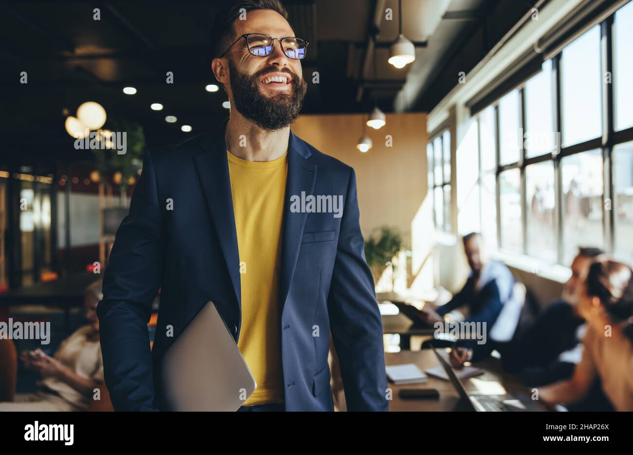 Felice uomo d'affari giovane che sorride in uno spazio di co-lavoro. Un uomo d'affari contemplativo che guarda via pensieroso mentre tiene un laptop. Allegro giovane entr Foto Stock