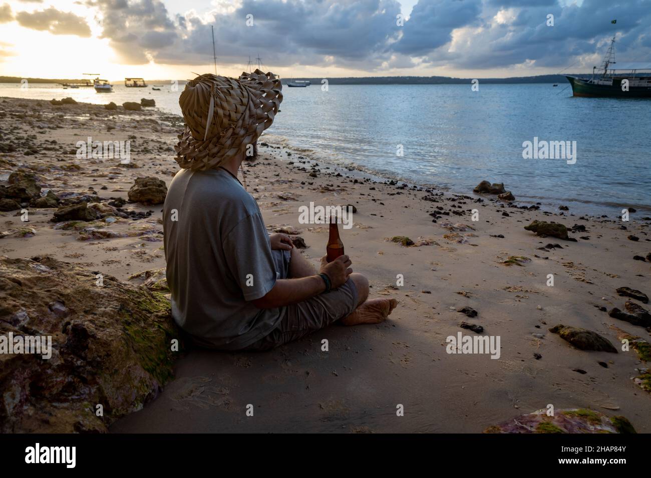 Uomo adulto con l'aspetto hippy o hippie avere una birra sulla spiaggia durante il tramonto. Il viaggiatore che ama il momento. Foto Stock