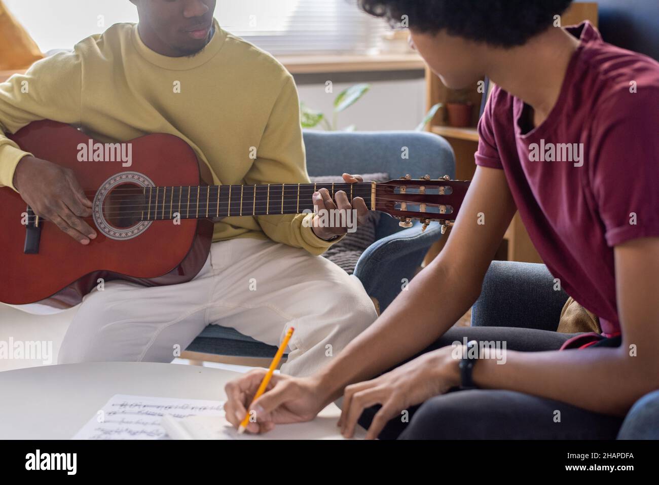 Giovane ragazzo biraciale che fa appunti e guarda l'insegnante di musica che suona la chitarra durante la lezione in ambiente domestico Foto Stock