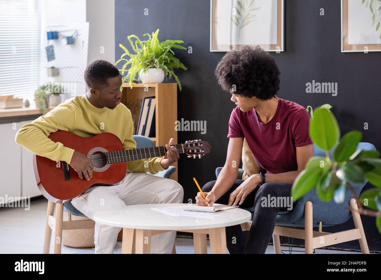 Insegnante di musica che suona la chitarra di fronte al suo studente seduto al tavolo e facendo note a lezione in ambiente domestico Foto Stock