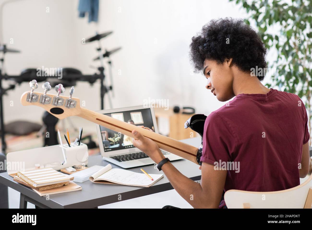 Giovane uomo seduto da una scrivania e che suona la chitarra davanti al computer portatile con il suo insegnante di musica sullo schermo Foto Stock