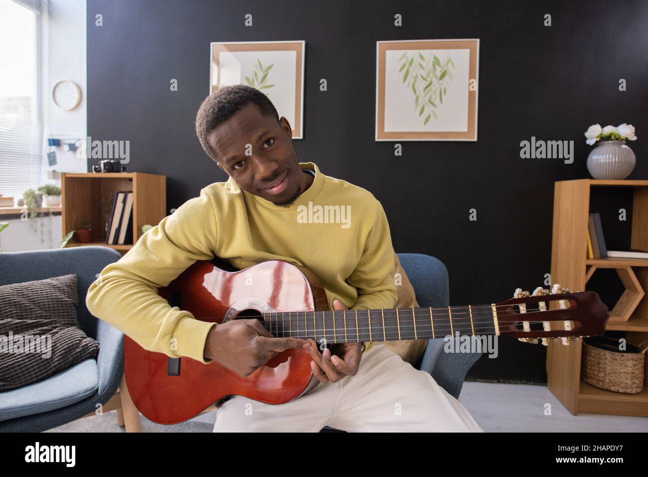 Giovane uomo africano sorridente con chitarra acustica seduto davanti alla macchina fotografica e guardato contro l'ambiente domestico Foto Stock