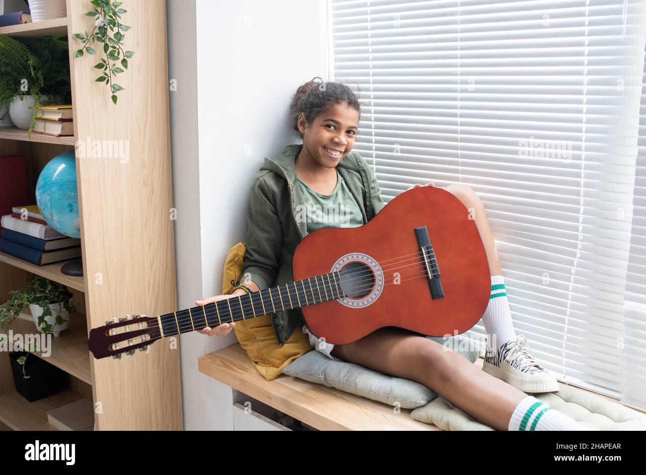 Felice ragazza biraciale carina con chitarra seduta sul davanzale e guardando la macchina fotografica in ambiente domestico Foto Stock