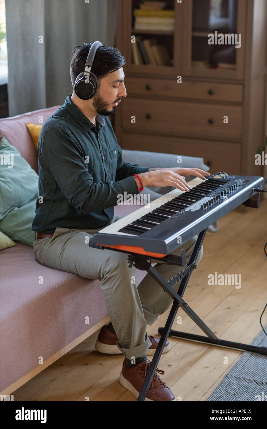 Vista laterale del giovane insegnante di musica in cuffie seduti sul divano con tastiera a piano e premendo i tasti Foto Stock