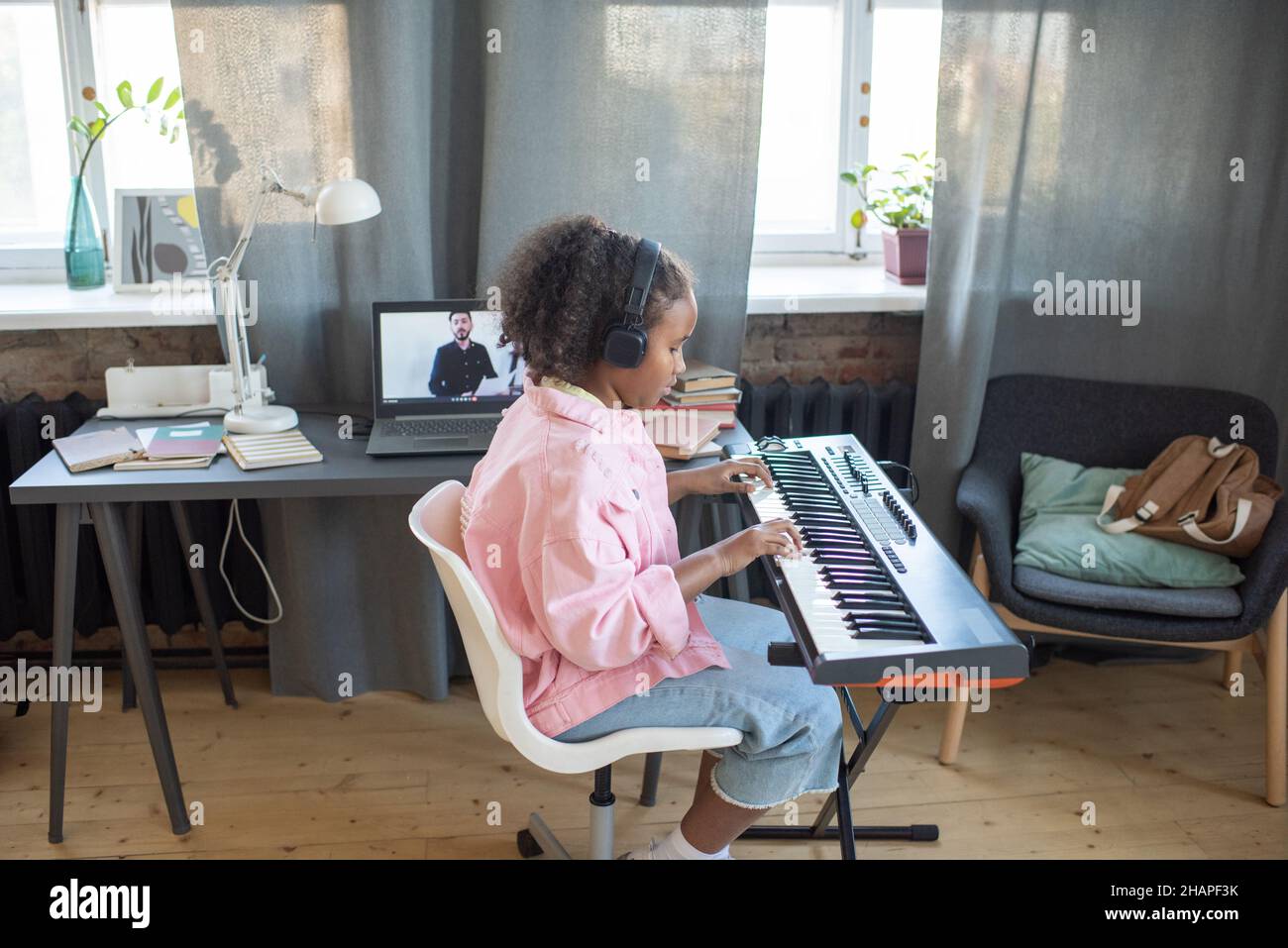 Vista laterale di una scolastica diligente che suona la tastiera del pianoforte mentre si siede contro la scrivania con il laptop durante la lezione online Foto Stock