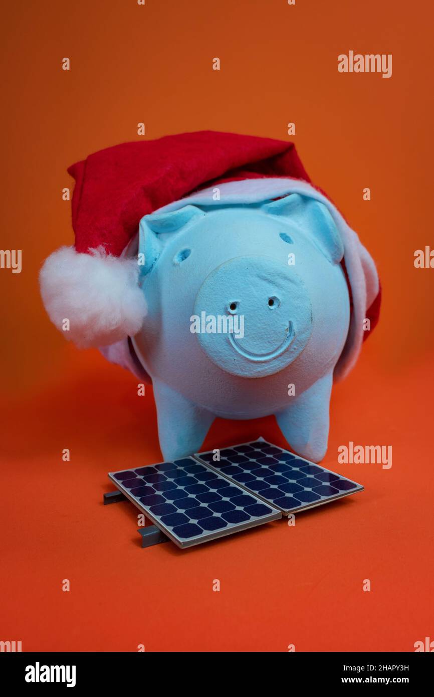 Monete di banca di Piggy con cappello di natale di Babbo Natale, pannello solare fotovoltaico e lampada isolato su sfondo arancione. Foto Stock