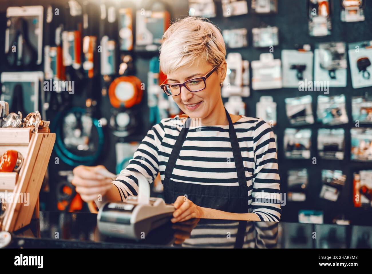 Sorridente lavoratore caucasico con capelli biondi corti e occhiali utilizzando il registratore di cassa mentre si trova in negozio di biciclette. Foto Stock