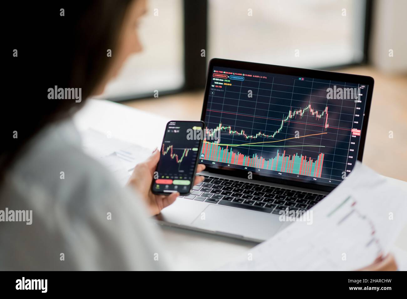 Un investitore e un operatore di crittografia di successo, utilizzando computer portatile e smartphone, analizza le carte di trading sul mercato azionario e lo scambio di criptovaluta digitale, conduce analisi, trading cripto monete Foto Stock