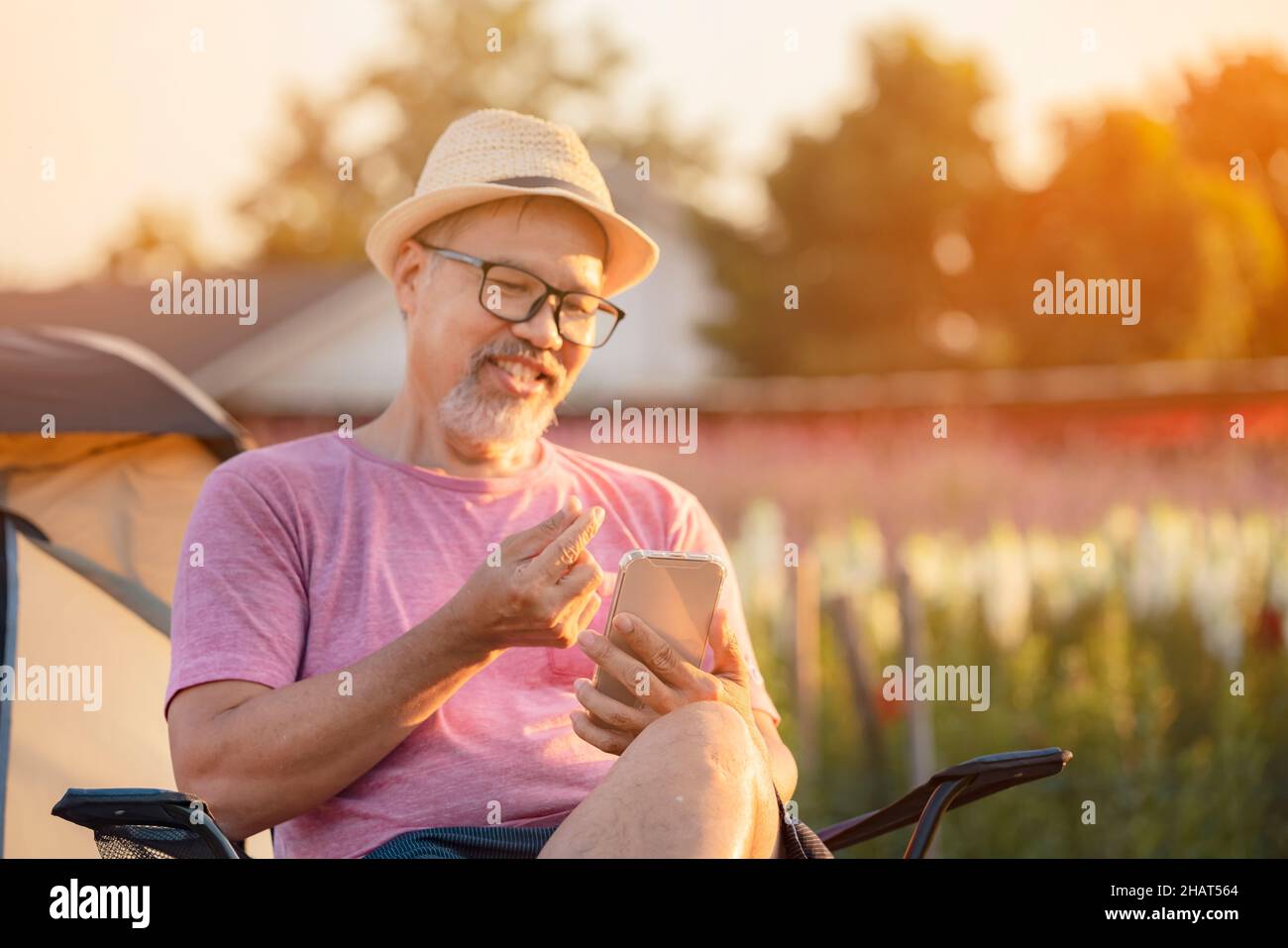 Focus su Telefono. Sorridente Asian Grey Beard Mate Man sedersi nella fattoria di fiori all'aperto e chitchat on online dating app nel telefono cellulare. Buon pensirreme Foto Stock