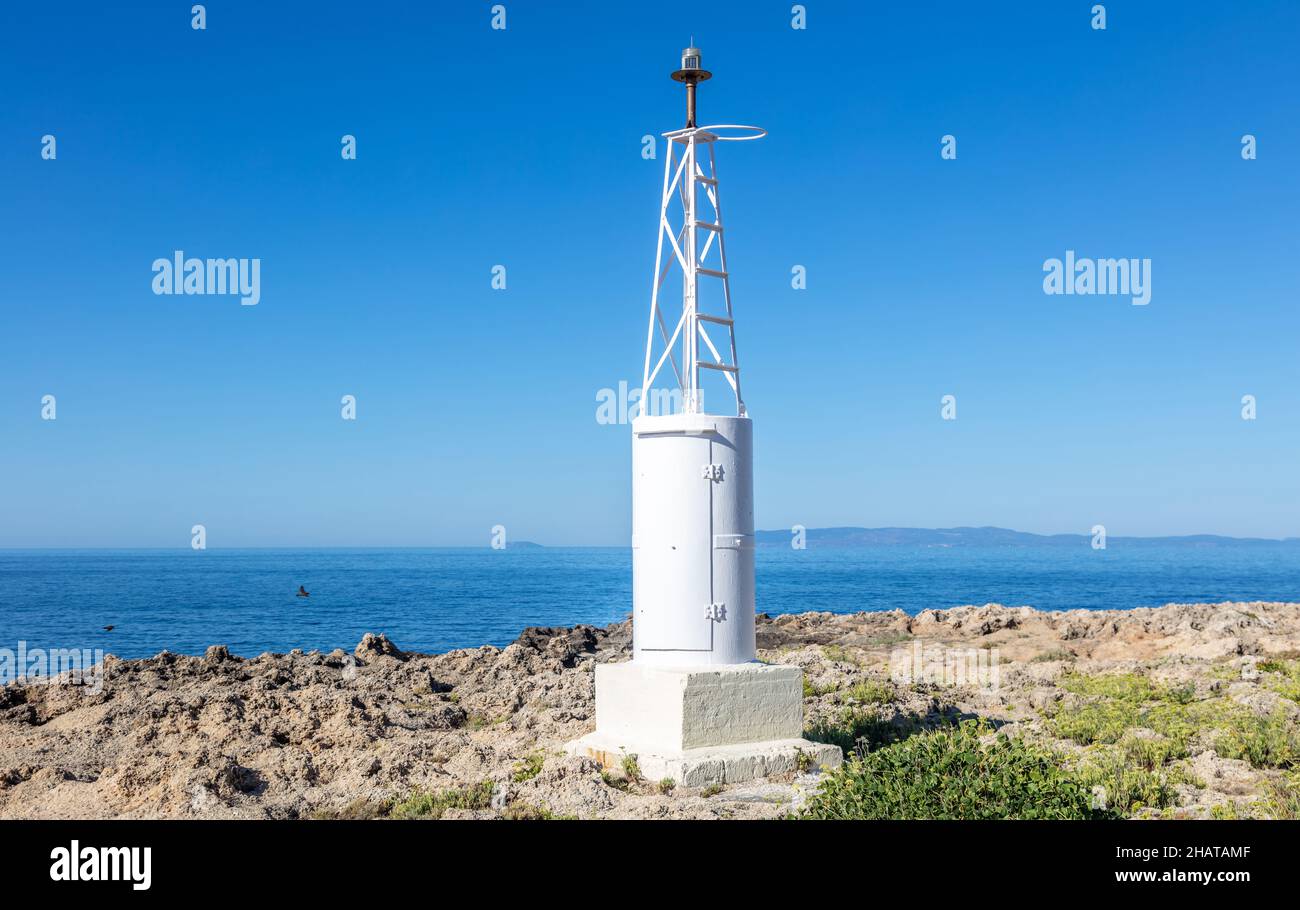 Safe navigation concept. Small lighthouse metal white construction at seaside for transport facilitate. Rocky land beacon, clear blue sky, calm Aegean Foto Stock