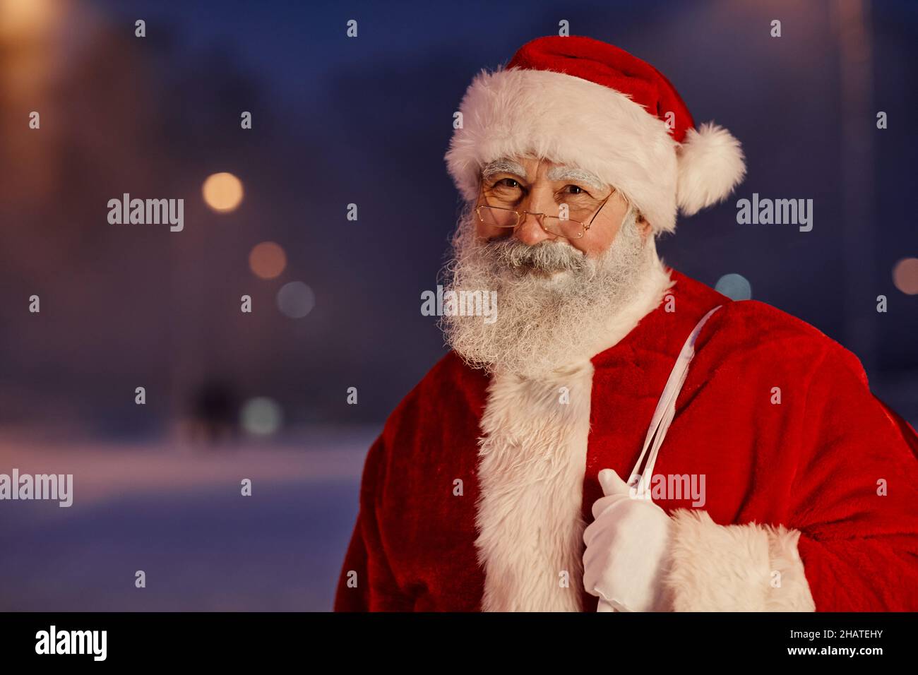 Ritratto orizzontale medio in primo piano di allegro Babbo Natale indossando abiti bianchi e rossi con occhiali in piedi all'aperto in serata guardando il camer Foto Stock