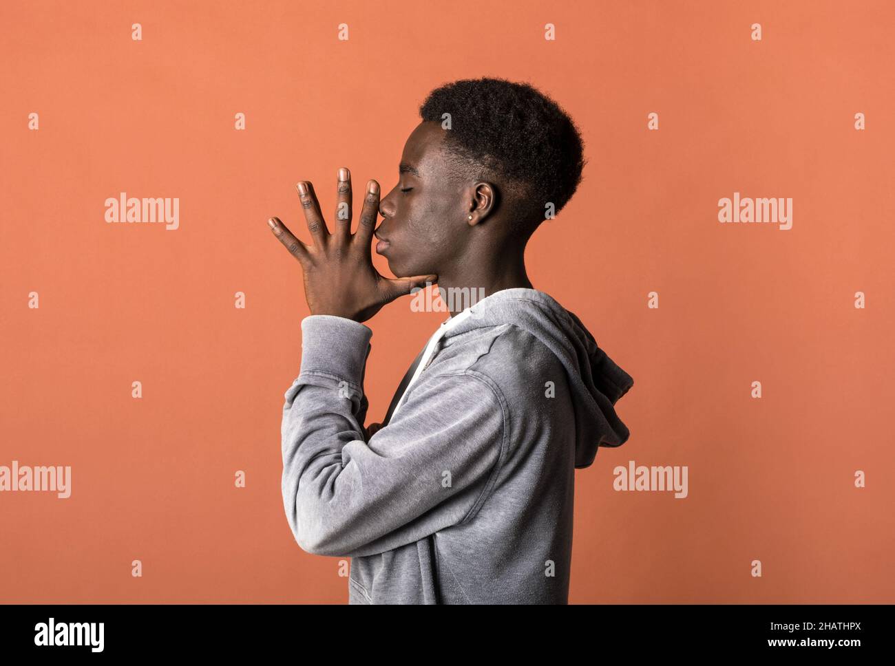 Vista laterale del giovane modello maschile africano americano con capelli ricci scuri in felpa con cappuccio grigio su sfondo arancione con occhi chiusi e. Foto Stock