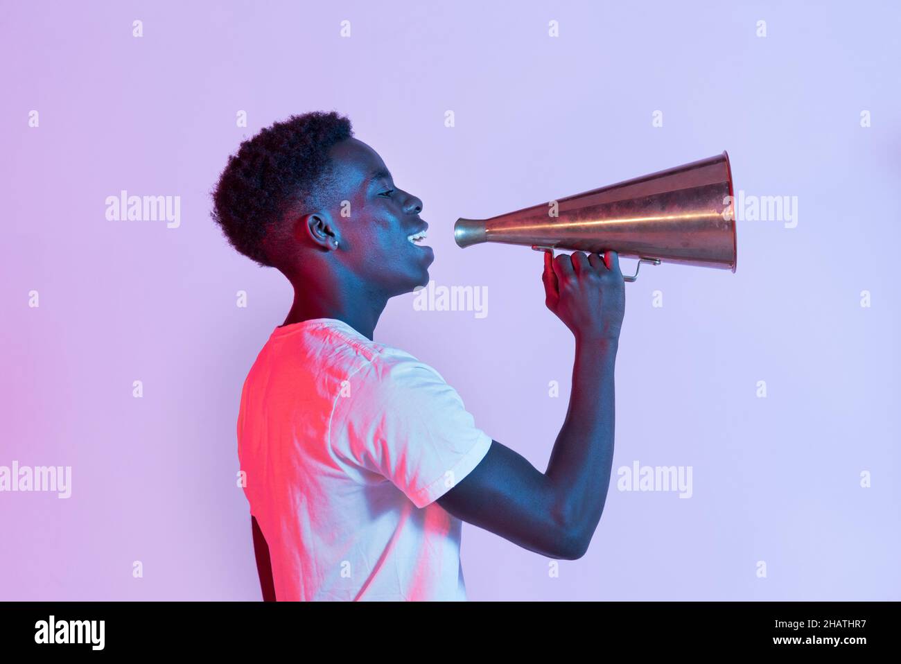Vista laterale di allegro giovane nero maschio millennial in t shirt sorridente mentre si parla con metallo vintage megaphone su sfondo viola Foto Stock