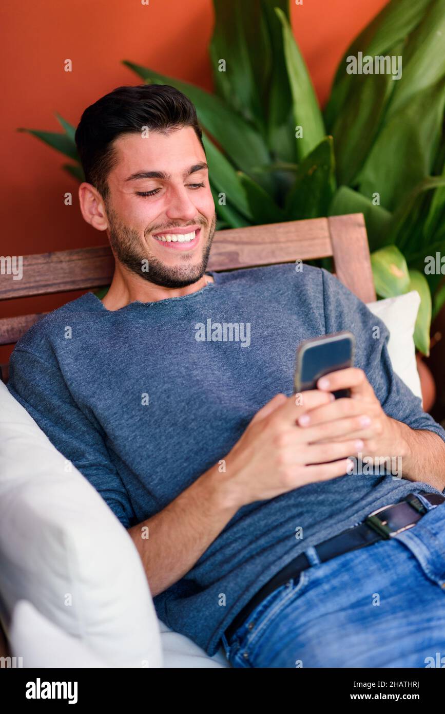 Messaggi di testo positivi bearded maschio sul cellulare mentre si siede in poltrona di legno con cuscino in camera con vaso verde pianta Foto Stock