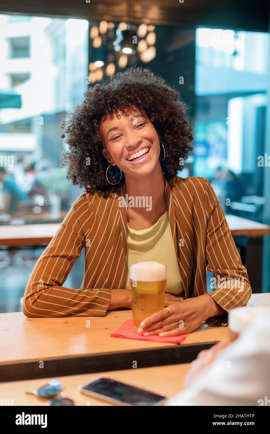 Allegra femmina afroamericana con capelli corti ricci in abiti casual seduti al banco bar in legno con un bicchiere di birra e ridendo al pub al giorno Foto Stock