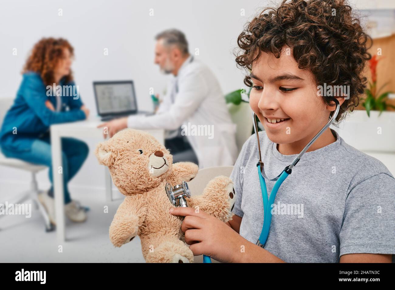 Bambino maschio felice che ascolta il suo peluche giocattolo orso usando stetoscopio in piedi nell'ufficio del medico con sua madre durante la consultazione pediatra Foto Stock