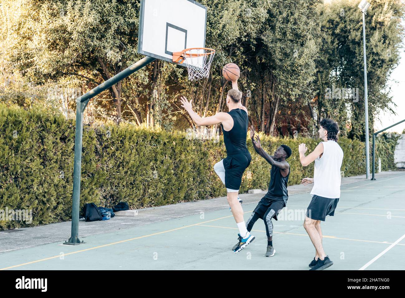 Uomo alto che salta per lanciare una palla in un cestino mentre gioca a basket con gli amici Foto Stock