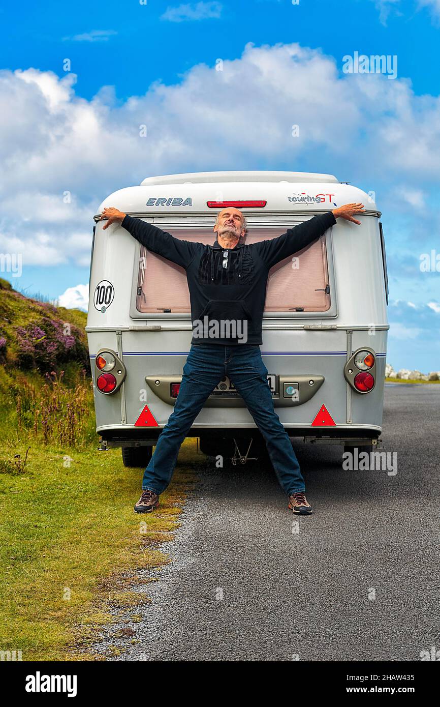 Uomo che si appoggia contro la roulotte in riva al mare, facendo una pausa, braccia e gambe formando una croce, strada panoramica Atlantic Drive, Achill Island, Mayo, Wild Foto Stock