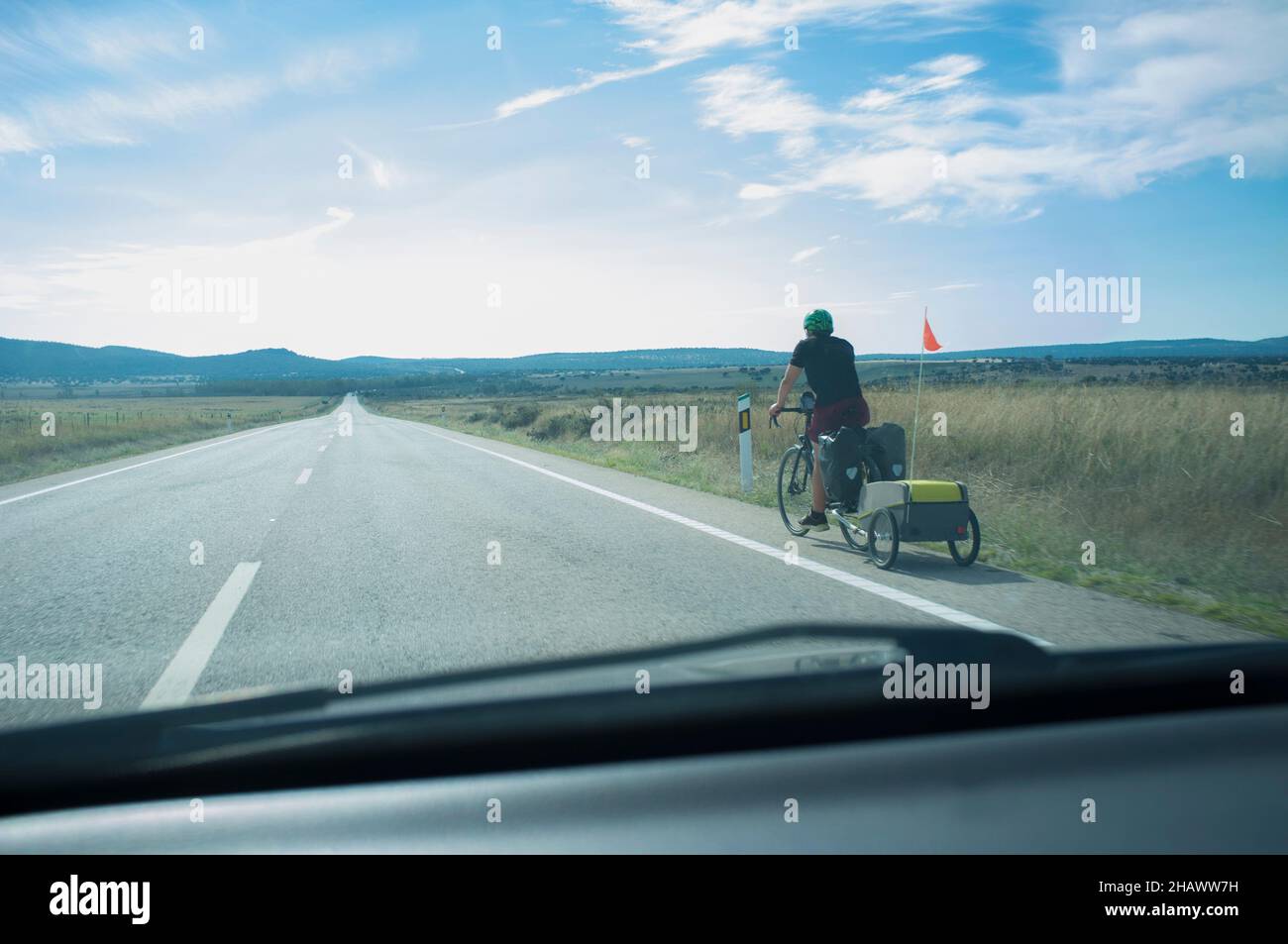 Biker sul percorso che tira il rimorchio da turismo della bicicletta. Strada di campagna con colline in fondo. Vista dall'interno della vettura Foto Stock