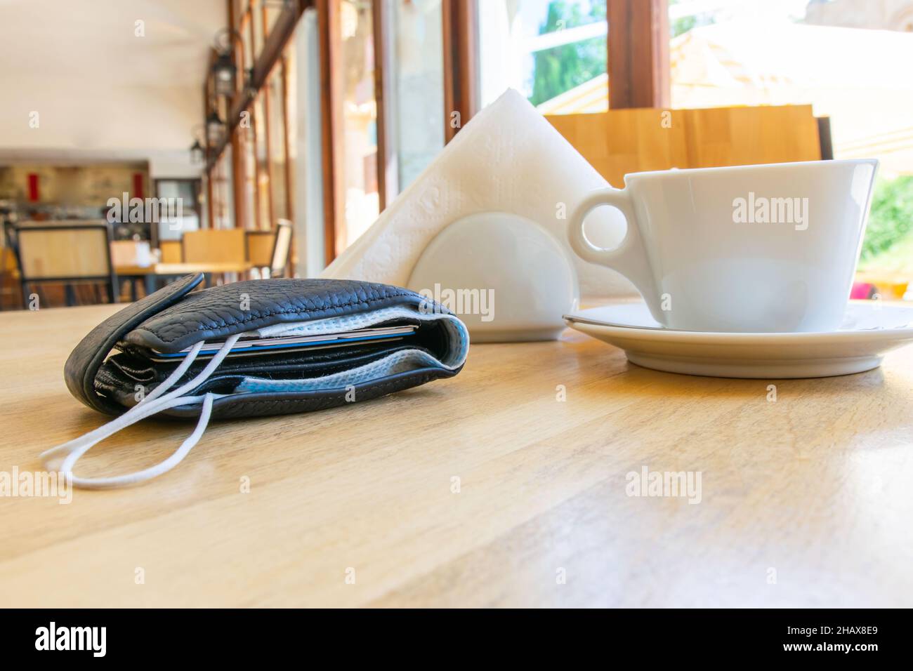 Portafoglio nero con maschera facciale all'interno con tazza bianca, tovaglioli in un supporto intorno in caffè senza persone. Norme e norme di sicurezza in post corona p Foto Stock