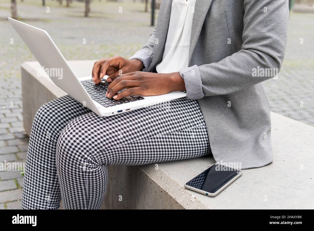 Ritagliare una donna d'affari afro-americana irriconoscibile digitando un messaggio e-mail sul computer portatile mentre si è seduti su una panca di pietra con lo smartphone nel parco Foto Stock