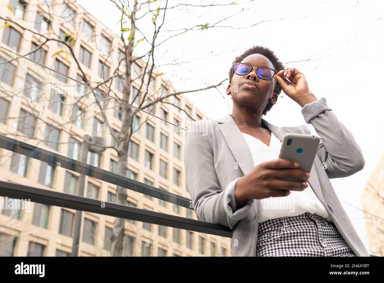 Dal basso del manager donna nero indossando abiti di classe browser smartphone mentre in piedi con gli occhi chiusi sul balcone con recinzione di vetro in ufficio buil Foto Stock