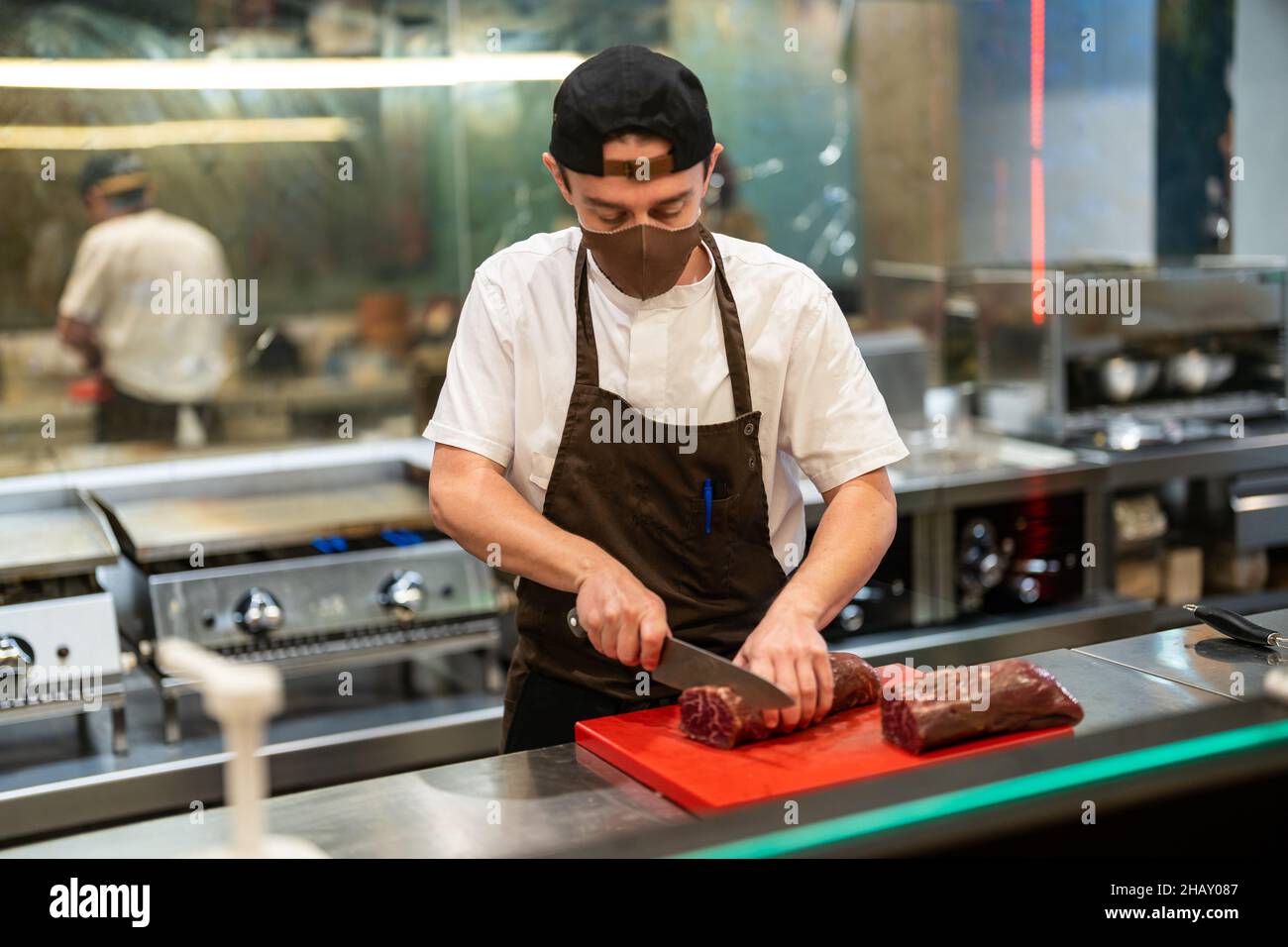 Attraverso un bicchiere di cuoco in grembiule tritare carne cruda a bordo mentre si lavora in cucina aperta del ristorante durante COVID 19 epidemia Foto Stock