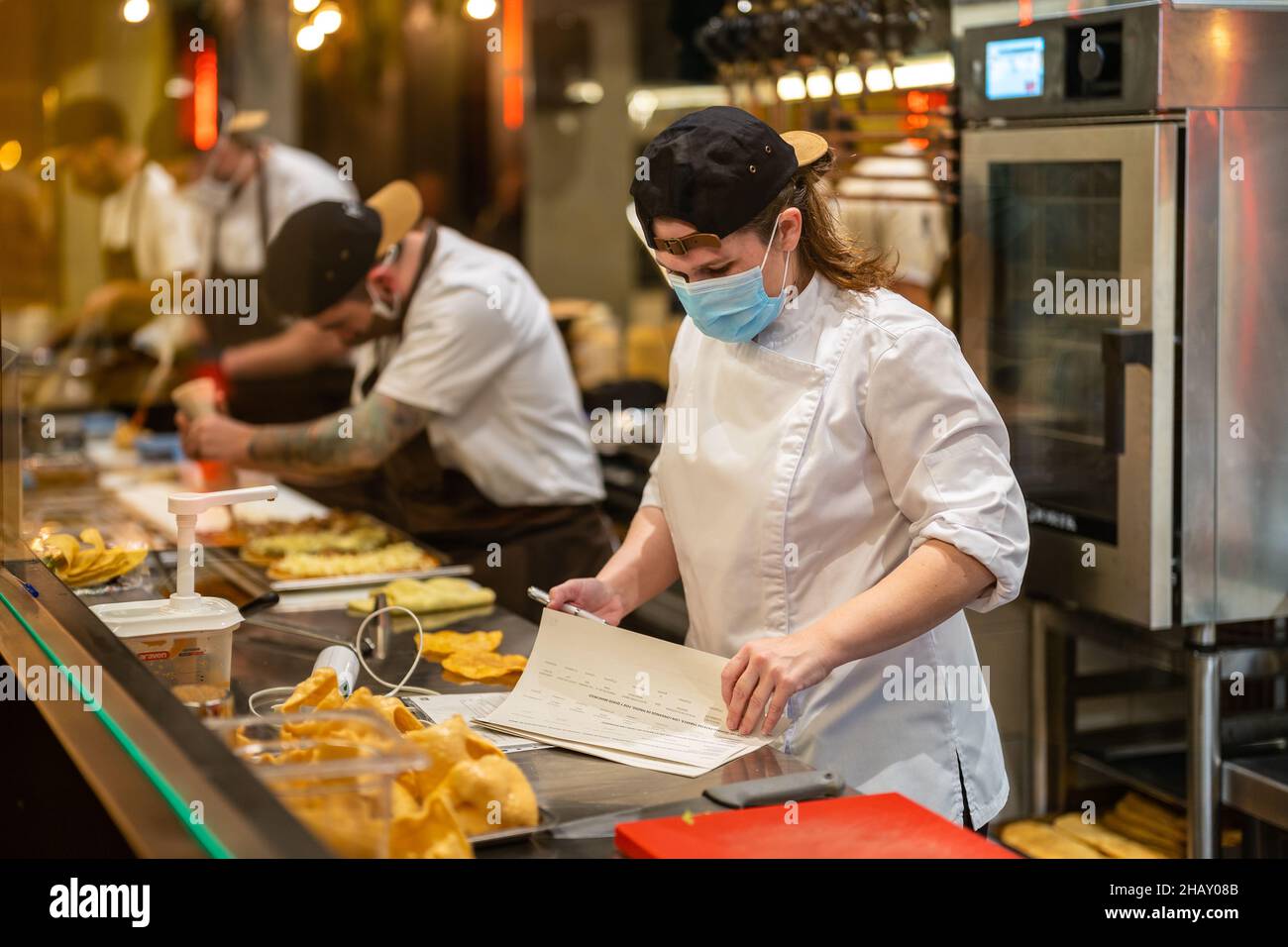 Attraverso un bicchiere di chef femminile indossando giacca bianca controllo fogli di carta mentre si trova al banco in cucina con cuochi impegnati a preparare il cibo durante il decor Foto Stock
