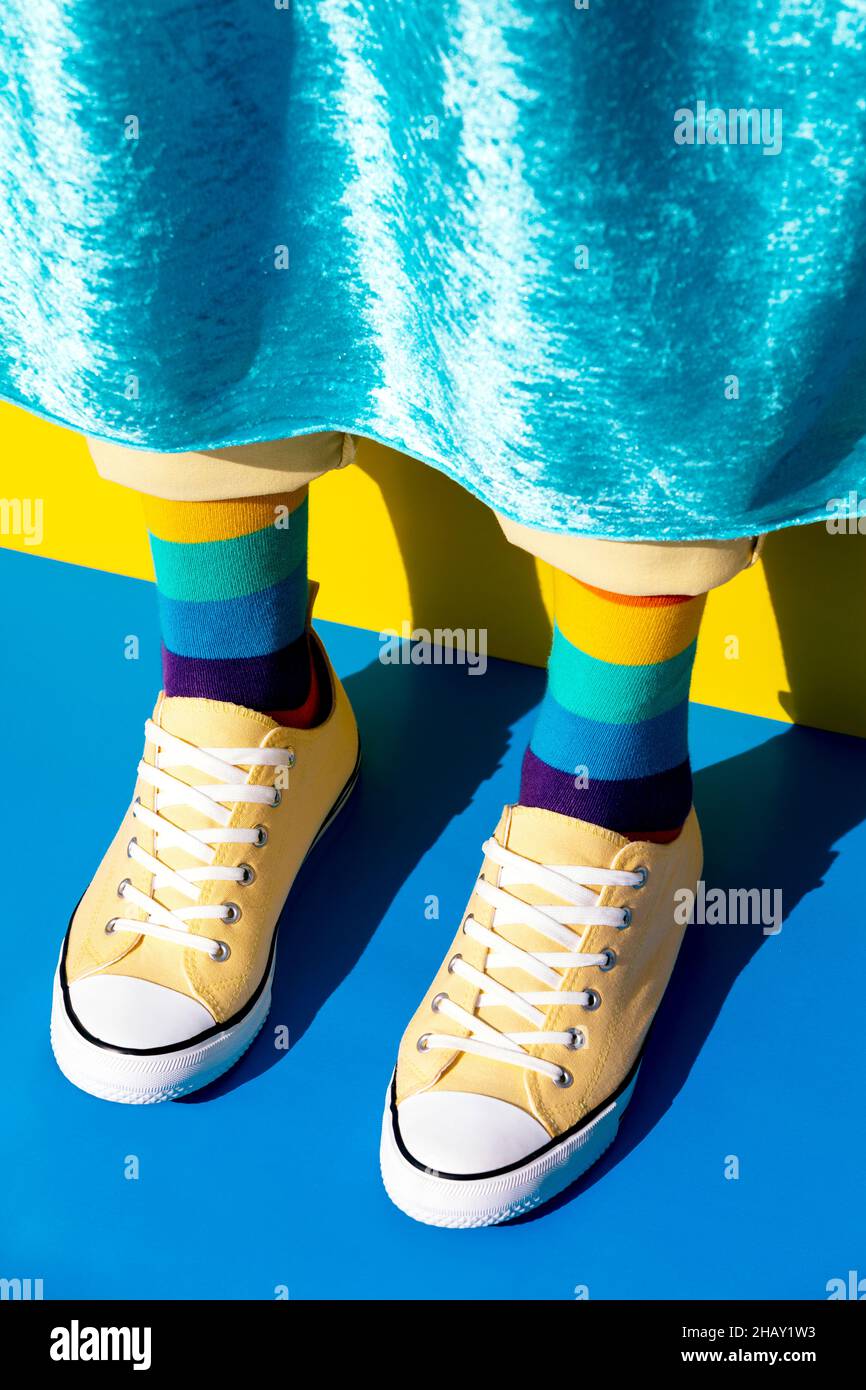 Crop anonymous omosessual persona indossare sneakers giallo e calze luminose con arcobaleno colori in piedi su superficie blu in camera luminosa Foto Stock