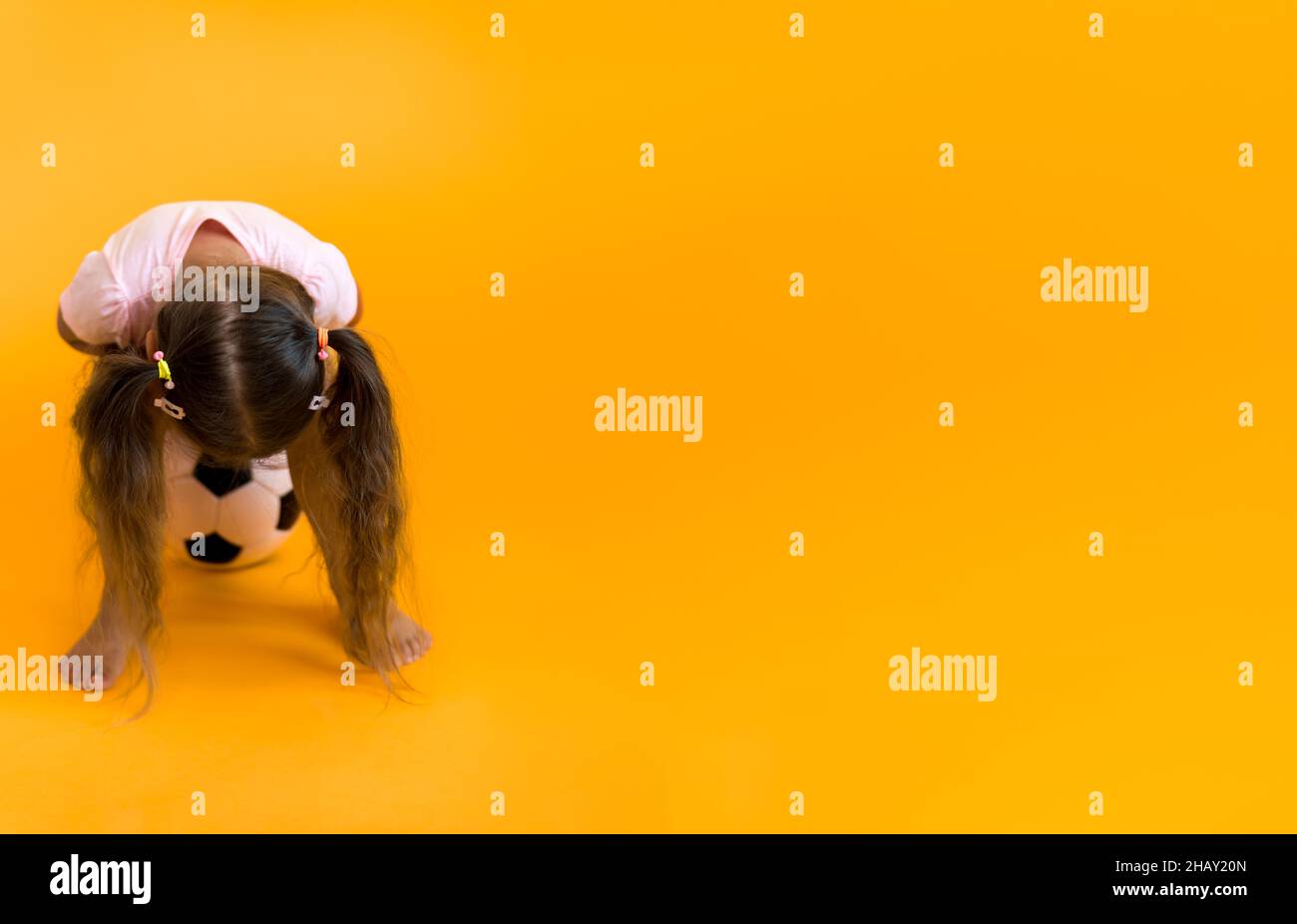 Autentica ragazza sorridente sorridente preschool con classica palla di calcio in bianco e nero guardare la fotocamera su sfondo giallo. Bambino giocare a calcio in t Foto Stock