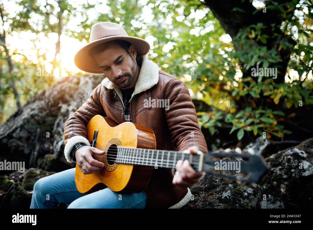 Musicista maschile talentuoso in cappello e outerwear che suona la canzone alla chitarra mentre si siede nella foresta vicino ad alti alberi verdi Foto Stock