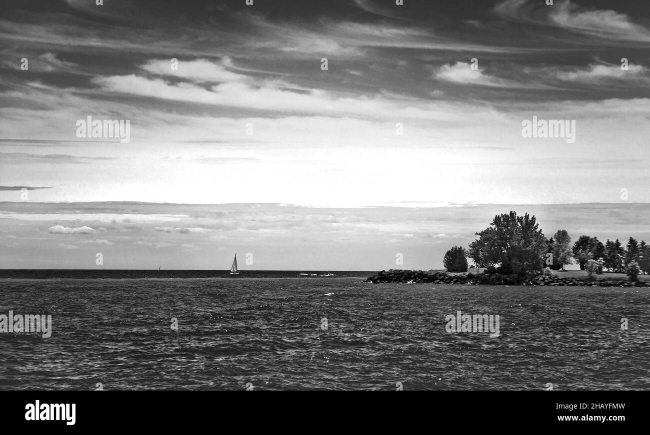 Bella stagcape con cielo nuvoloso, isola con alberi, e una piccola barca a vela che taglia le onde nel vento. Foto Stock