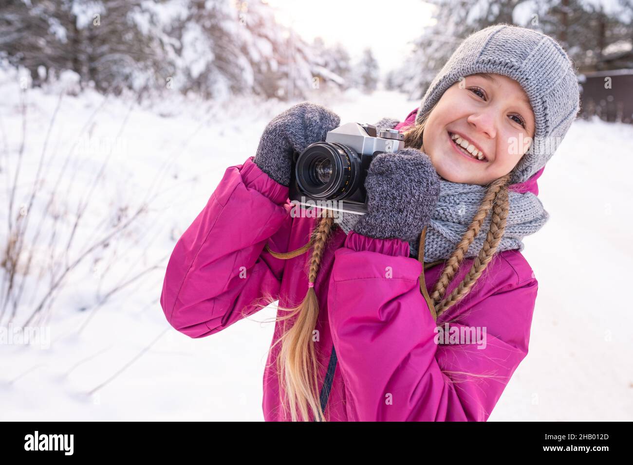 Ritratto di ragazza adolescente sorridente con lunghi pigtail in abiti caldi, tenendo la macchina fotografica in mani, contro alberi di pino nevoso. Bambini in inverno a piedi. Giorno di Foto Stock