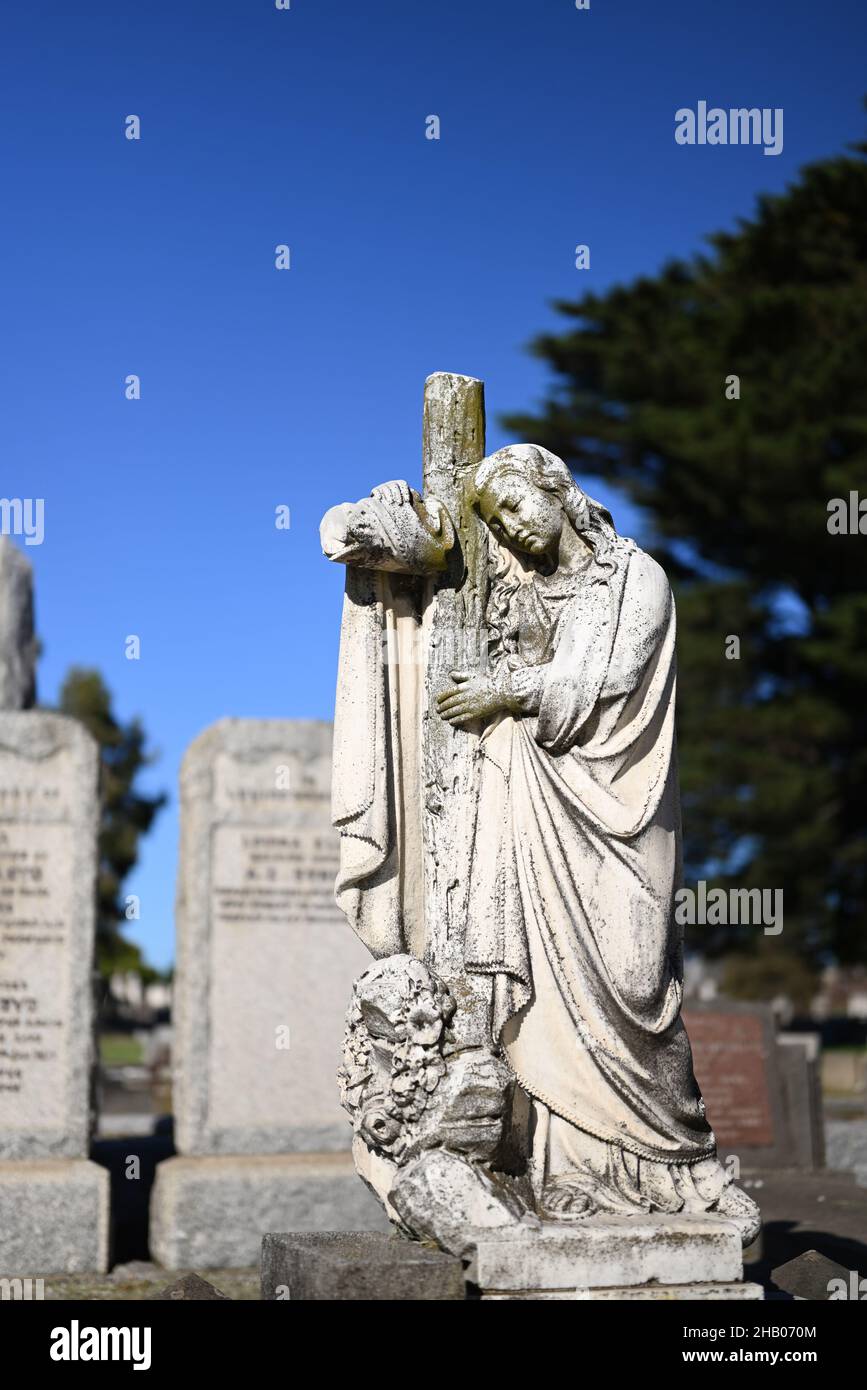 Ritratto di una scultura in pietra di una donna drappeggiato su una croce cristiana in un cimitero, con tombe sullo sfondo Foto Stock