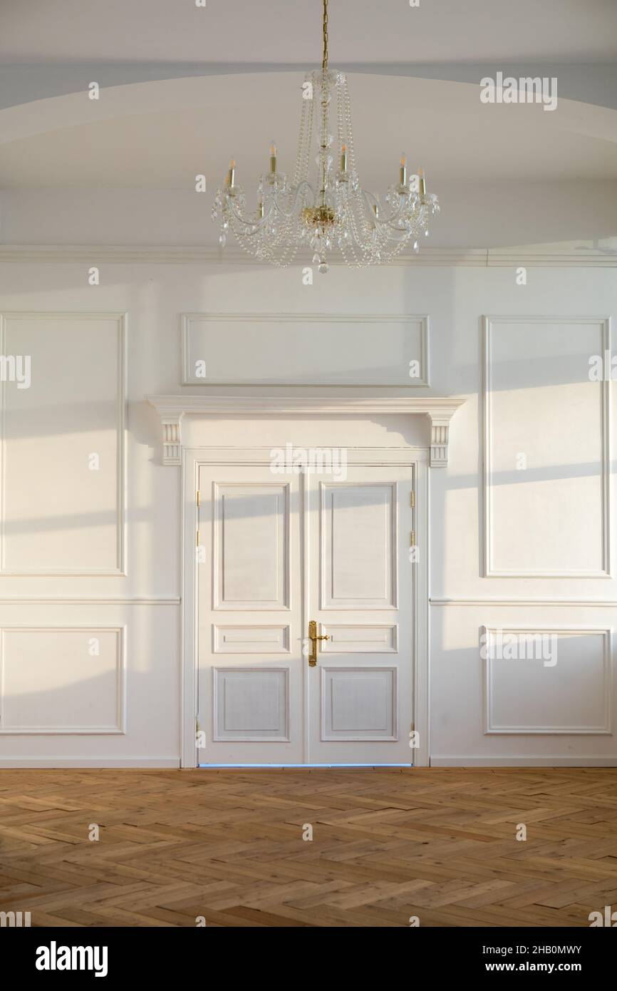 Porte chiuse bianche nella luce spaziosa stanza vuota con design classico e lampadario appeso al soffitto vicino alla parete con luce del sole Foto Stock