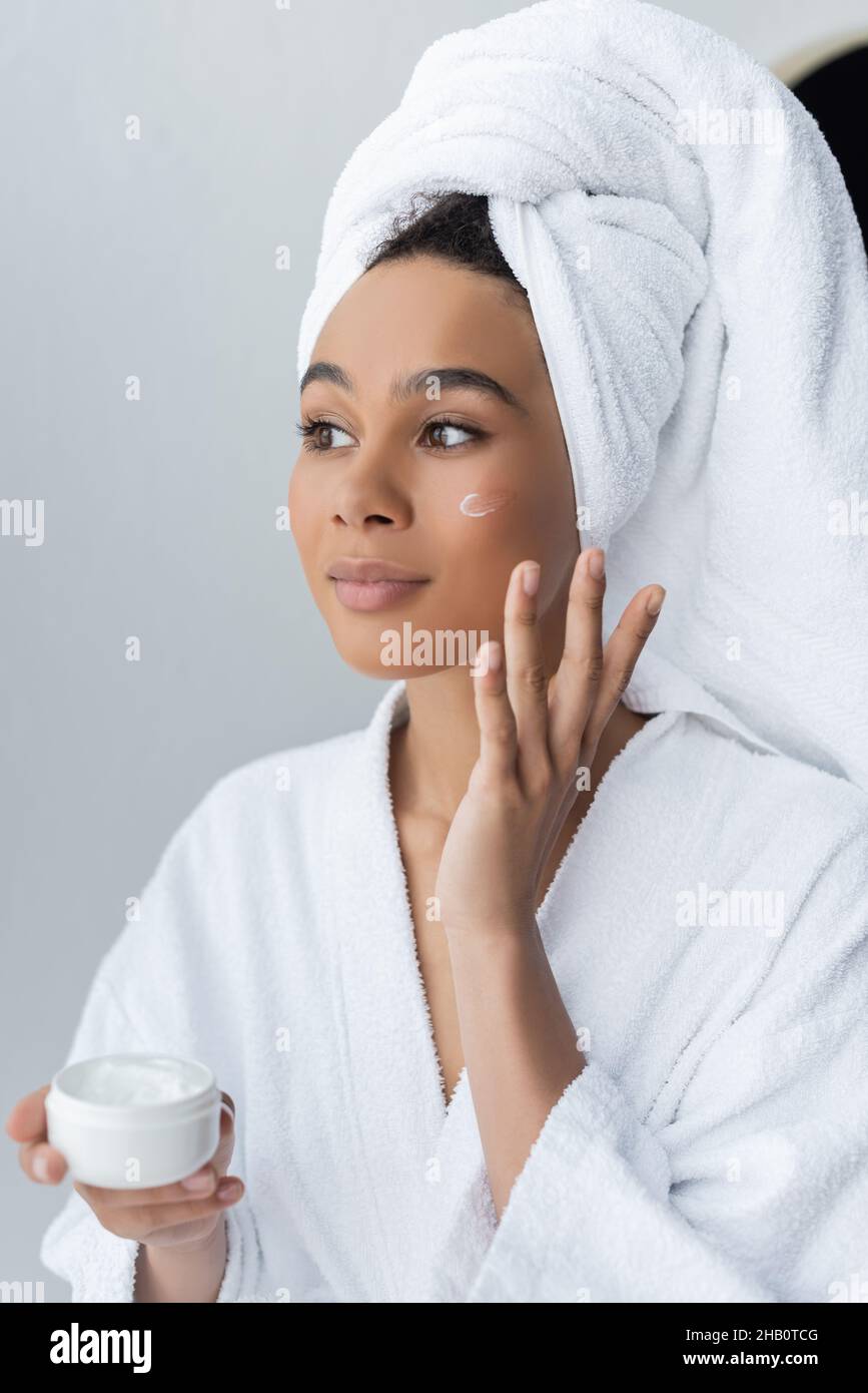 donna afro-americana da sogno in accappatoio contenitore con crema viso in bagno Foto Stock