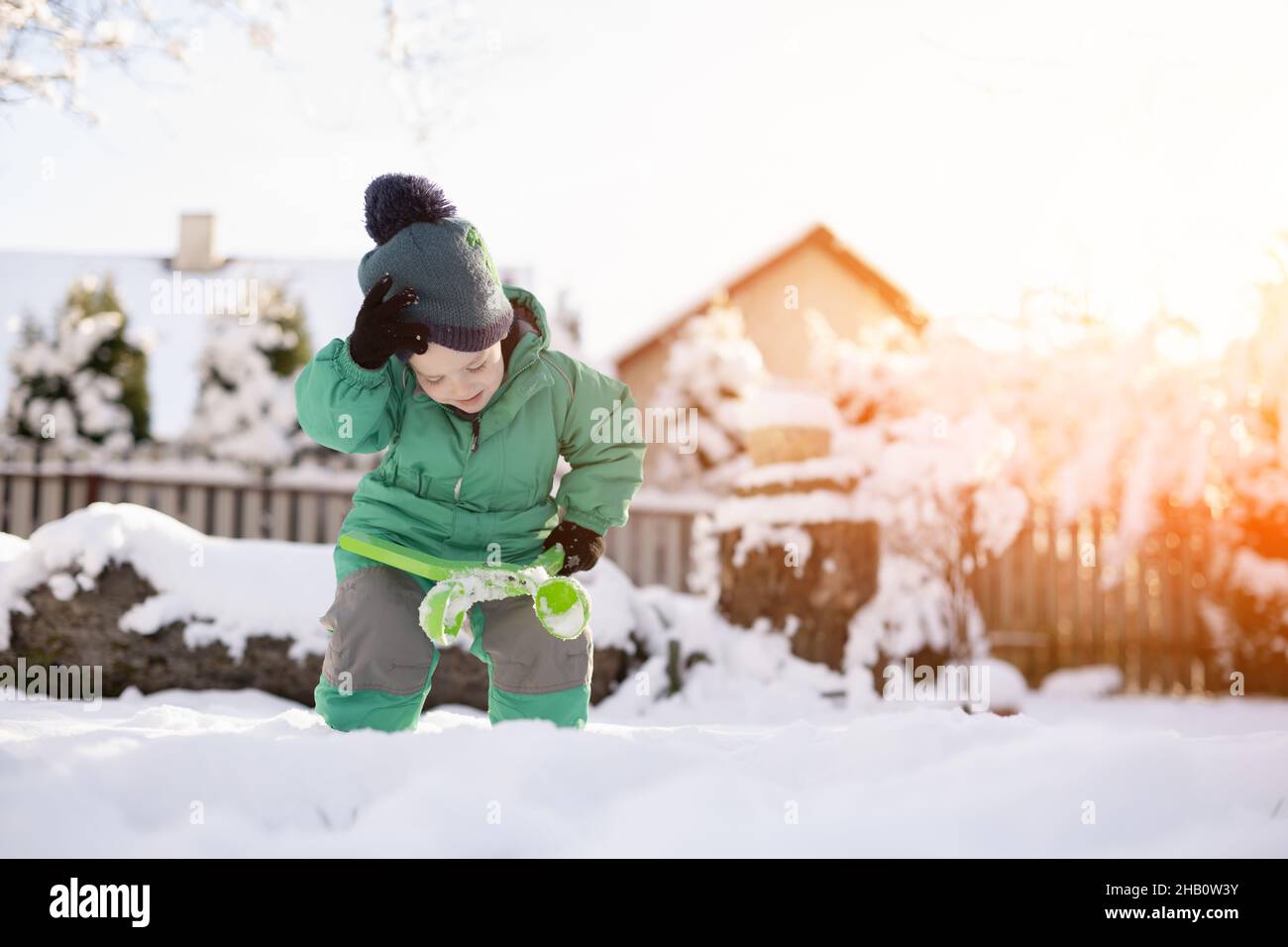 Ragazzo che gioca con la neve in inverno. Bambino piccolo in giacca verde e cappello a maglia fare le palle di neve vicino a casa a Natale. Giochi per bambini e divertimento nel giardino innevato Foto Stock