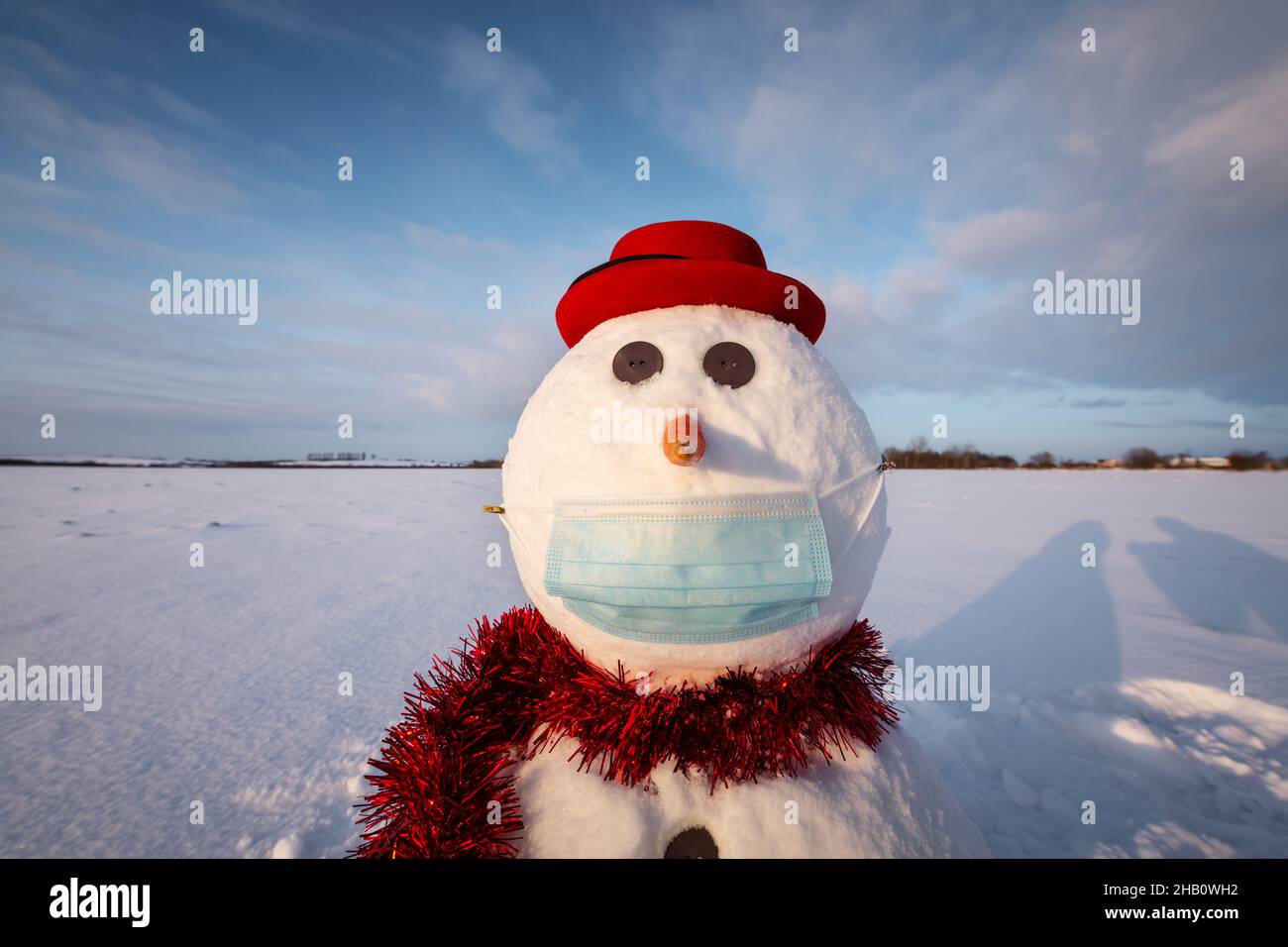 Simpatico pupazzo di neve in elegante cappello rosso con maschera medica su campo innevato. Concetto di allontanamento sociale e di viaggio annullato Foto Stock
