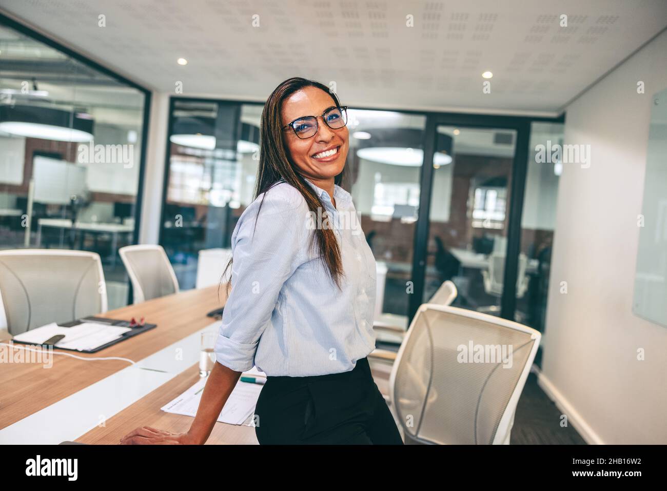 Donna d'affari di metà-adulto che sorride allegro in un ufficio. Donna d'affari felice appoggiata contro un tavolo in una sala riunioni. Abbigliamento allegro imprenditore femminile Foto Stock