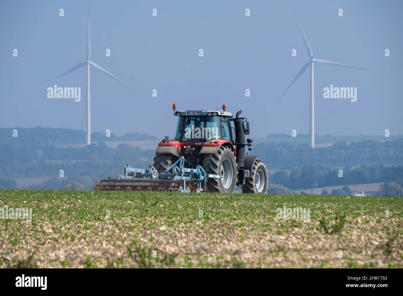 Trattore che gira su suolo e mulini a vento in distanza a Puyrolland (Francia centro-occidentale): Trattore con erpice in un campo Foto Stock