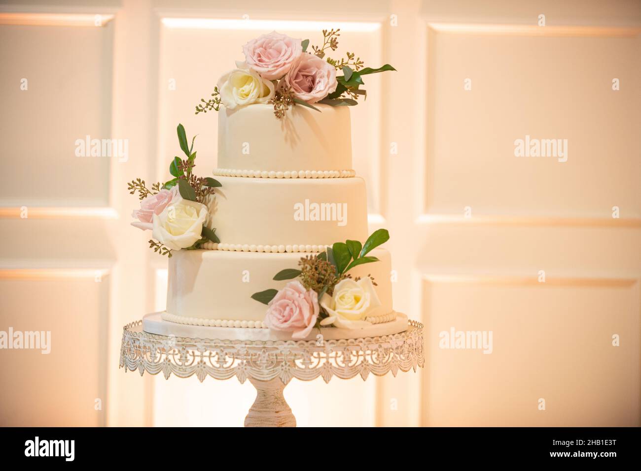 Splendida torta nuziale a tre livelli con perle e fiori rosa e bianchi sul vassoio torta in pizzo vicino alla parete bianca Foto Stock