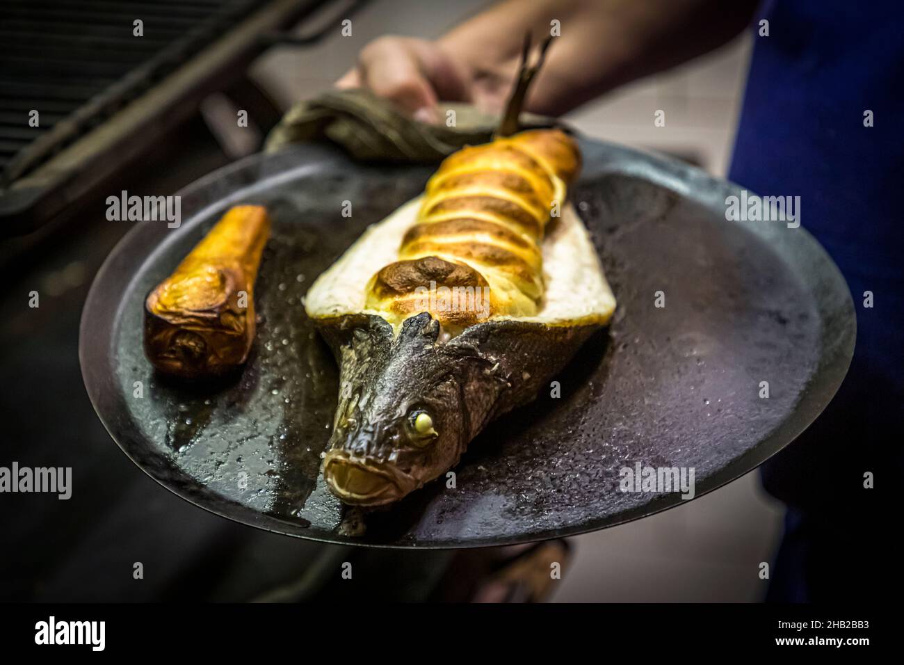 Un Loup de Méditerranée (branzino mediterraneo) ripieno di un sofflé delicatamente tartufato poggia su un piatto grill nero al Restaurant Hestia a Bormes-les-Mimosas, Francia; lo chef Cyril Aveline tiene il piatto appena uscito dalla griglia Josper, con un pezzo più piccolo arrosto accanto. Foto Stock