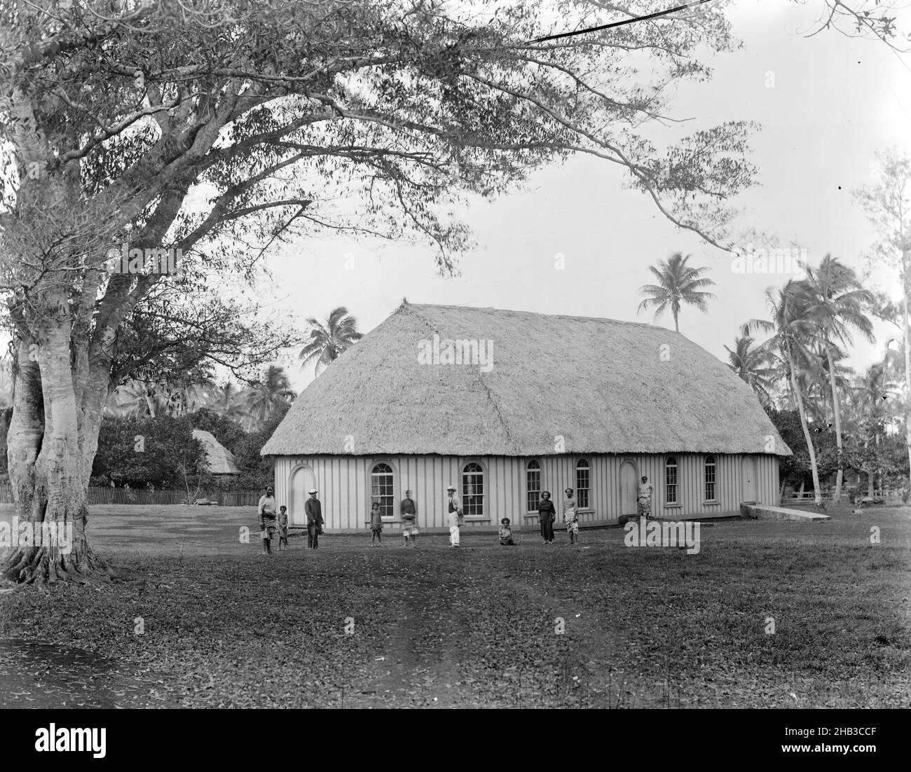 Neiafu, Wesleyan Church (esterno), Burton Brothers studio, studio fotografico, 26 luglio 1884, Nuova Zelanda, fotografia in bianco e nero, grande albero lasciato in primo piano nella radura di fronte alla chiesa edificio. Dieci persone in piedi o seduti sono di fronte alla chiesa. La chiesa ha un tradizionale tetto di favola, con pareti in legno, porte e finestre in stile coloniale. Alti alberi di cocco sono sul retro della chiesa, c'è il tetto di un altro edificio visibile in fogliame tropicale direttamente dietro l'albero Foto Stock
