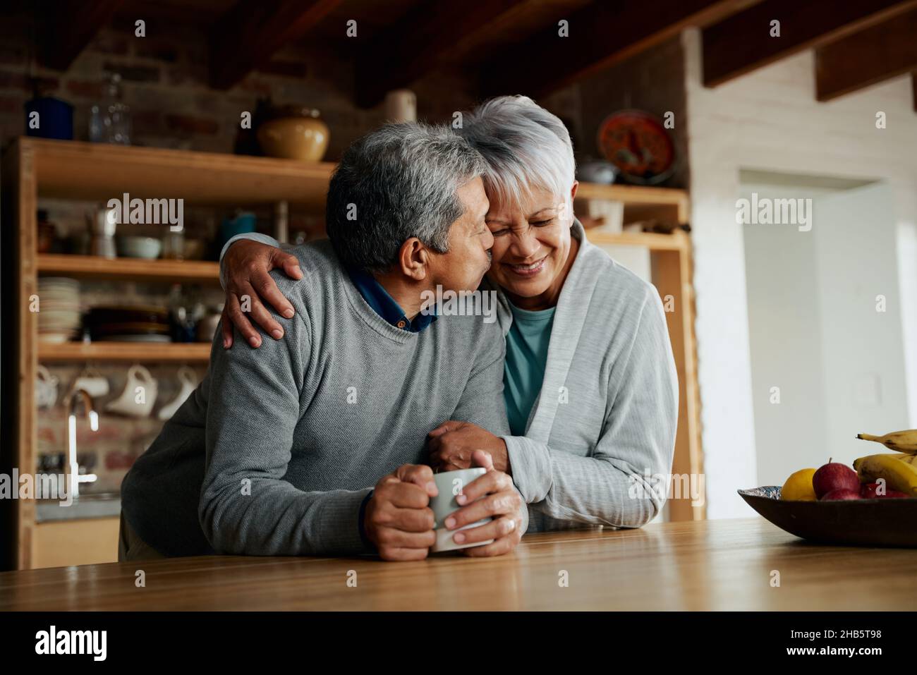 Felicemente anziana coppia biraciale che gode di pensione. Moglie che tiene marito in cucina moderna. Il marito bacia la moglie sulla guancia. Foto Stock