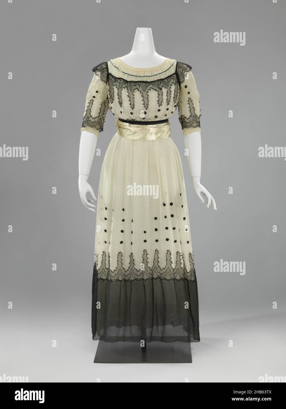 Abito a tre strati di georgette crespata, il camoscio è composto da tre strati sovrapposti di georgette crespata; un abito bianco, una abito 'eau de nil' e una abito bianco e nero. Questo ha dato al tutto un bagliore blu-verde. Sorprendentemente contemporanei sono i cerchi di nastro laminato in raso nero sulla gonna bianca e la frangia annodata nera e bianca sul nastro. La gonna è priva di piedi e abbastanza spaziosa per un'adeguata libertà di movimento., anonima, anonima, Paesi Bassi (possibilmente), Francia (possibilmente), c. 1916 - c. 1919, geheel: seta, pizzo, lunghezza 33,0 cm lunghezza 102,0 cm vita 28,0 cm Foto Stock