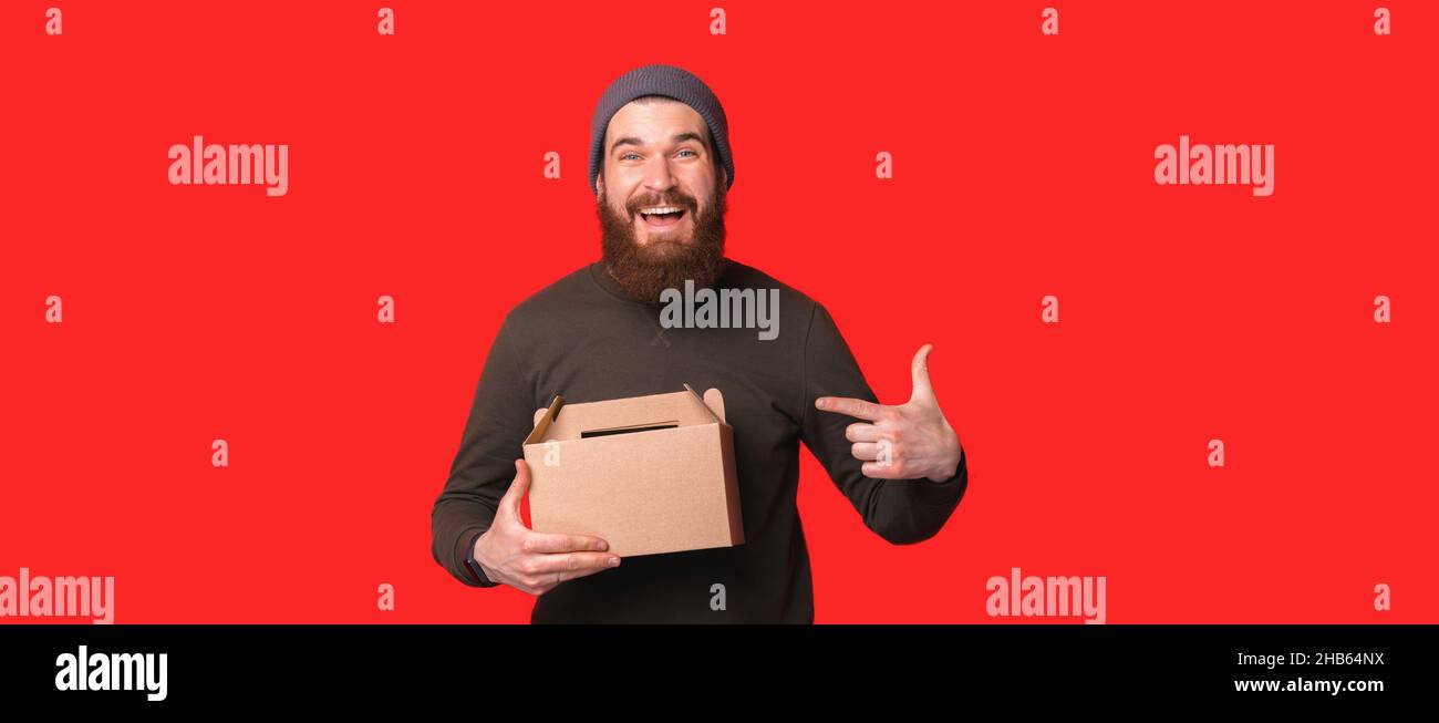 Il maschio allegro bearded punta alla scatola di cartone mentre raccomanda il pacchetto eco in studio contro la parete rossa e guardando la macchina fotografica Foto Stock